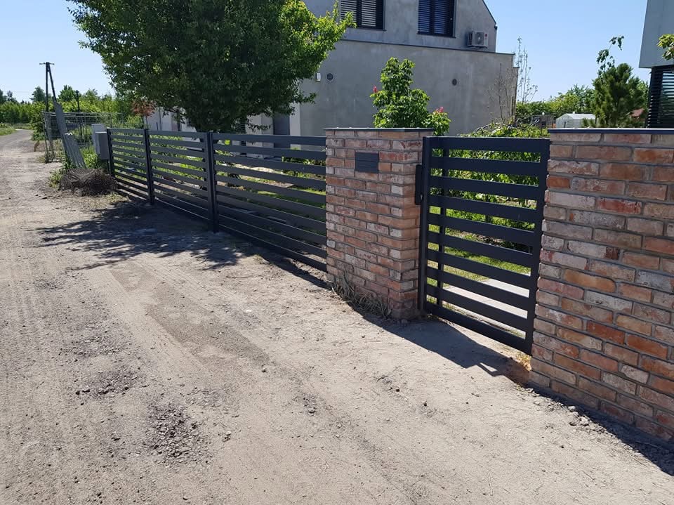 Nowoczesna, pozioma, metalowa brama wjazdowa w kolorze grafitowym, osadzona w murze z czerwonej cegły, prowadząca na żwirową drogę.