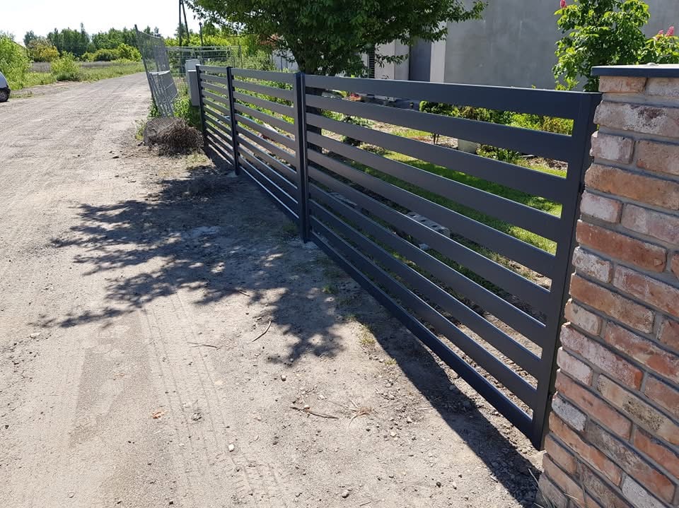 Nowoczesne, poziome przęsła ogrodzenia metalowego w kolorze grafitowym, osadzone na słupkach z cegły klinkierowej, z widocznym fragmentem drogi gruntowej i cieniem drzew.