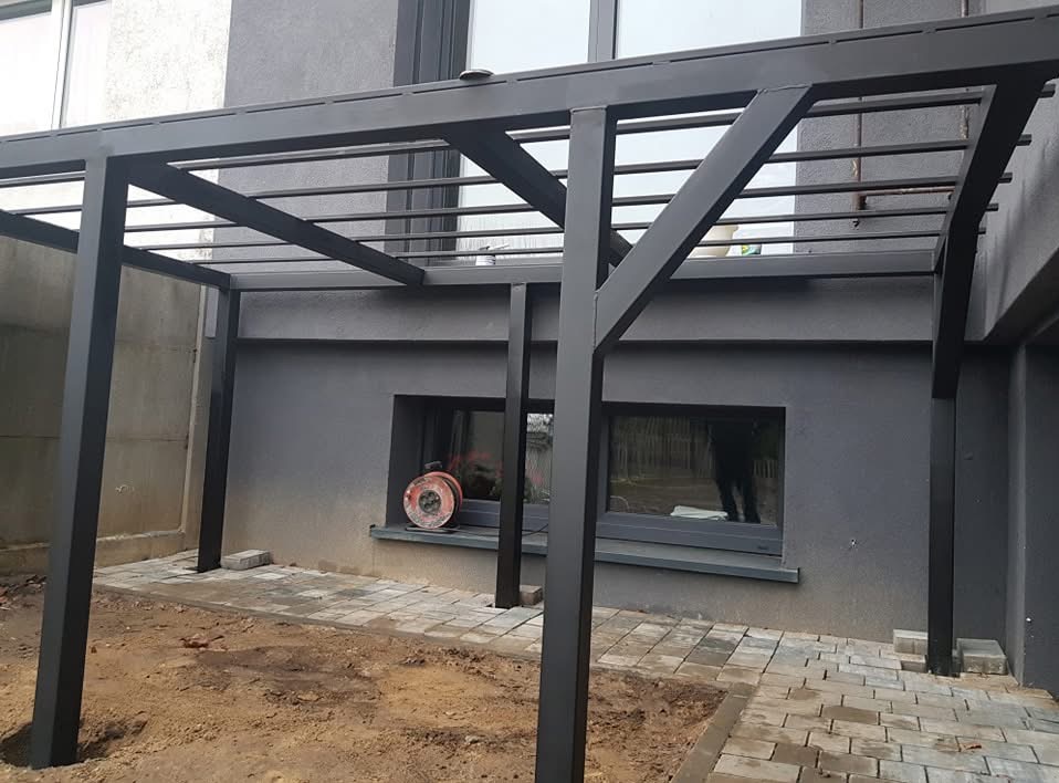 Czarna metalowa pergola z poziomymi belkami, częściowo wyłożona kostką brukową, z widocznym zwiniętym kablem na parapecie okna w tle.