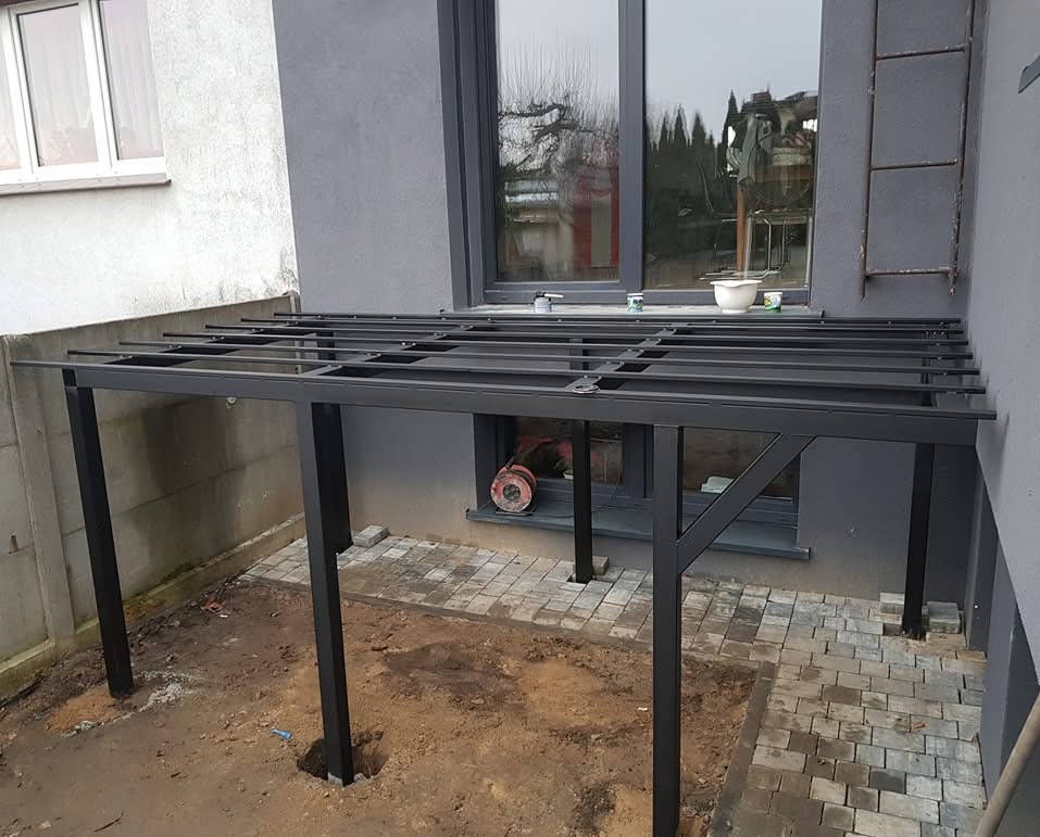 Montaż czarnej metalowej pergoli przy szarym domu, widoczne fundamenty i częściowo ułożona kostka brukowa.