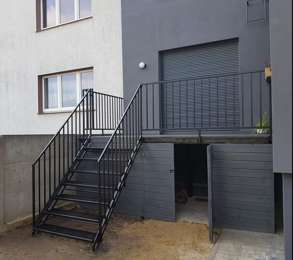 Zewnętrzne schody metalowe z czarną balustradą prowadzące do podwyższonego tarasu z szarą roletą i wejściem do garażu, widok na dwa budynki o różnej kolorystyce elewacji.