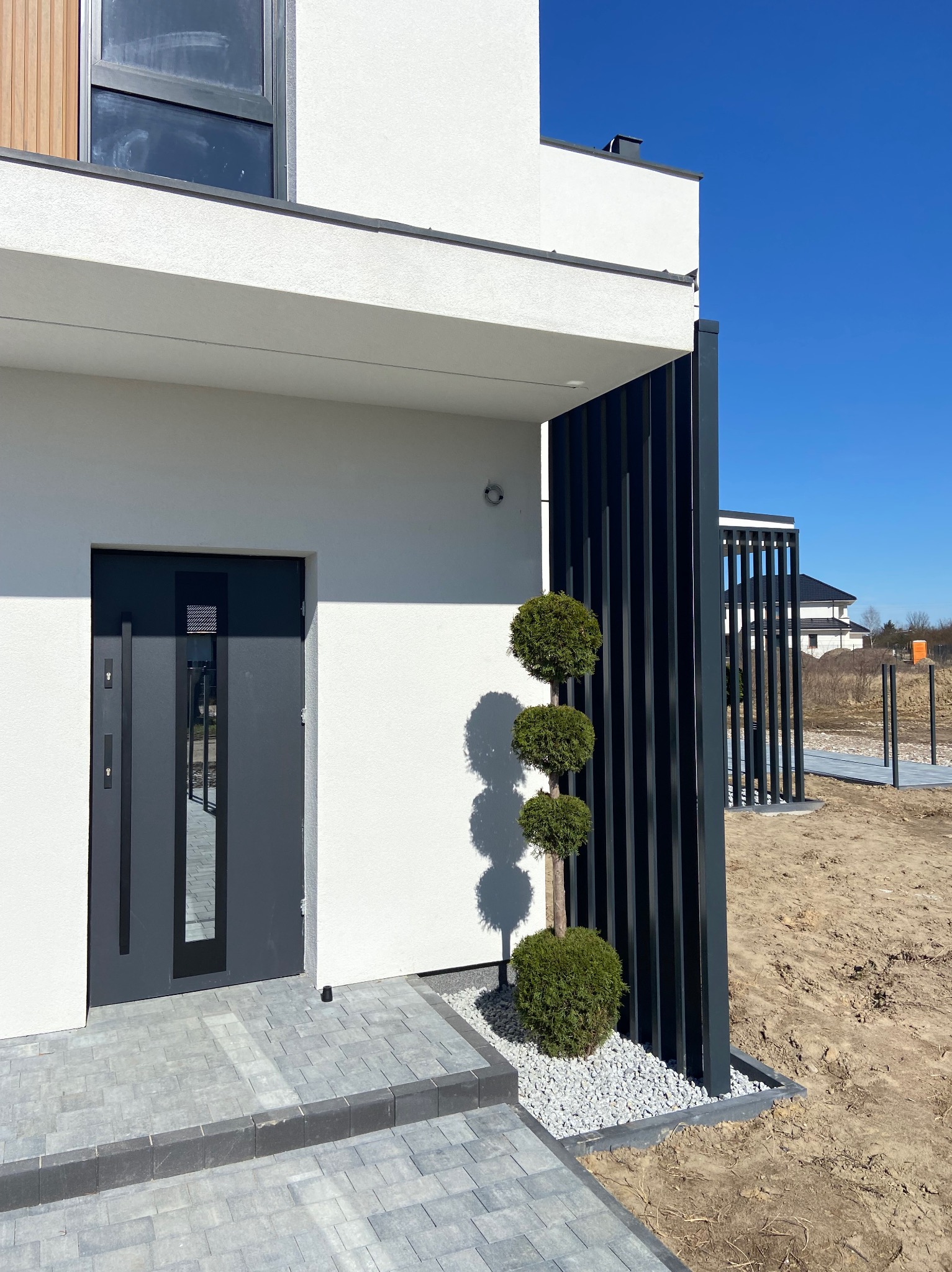 Nowoczesny dom jednorodzinny z antracytowymi elementami: drzwi wejściowe, pionowe panele dekoracyjne oraz kuliste krzewy na tle białej elewacji i kostki brukowej.