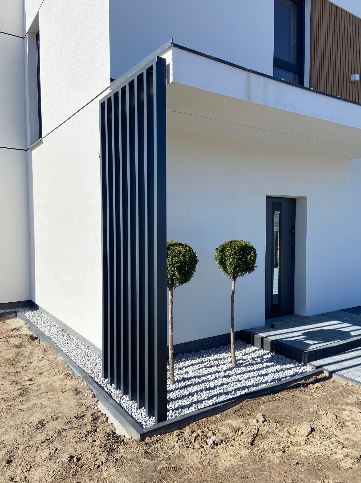 Nowoczesna fasada domu z pionową, metalową konstrukcją w kolorze antracytowym, ozdobnymi kulistymi drzewkami i szarą, kamienną rabatą, widok na wejście do domu.