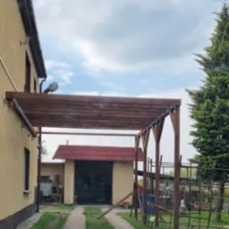 Konstrukcja z ciemnego drewna przymocowana do ściany domu, prawdopodobnie w trakcie budowy, z widocznymi słupami i belkami na tle budynku gospodarczego i pochmurnego nieba.