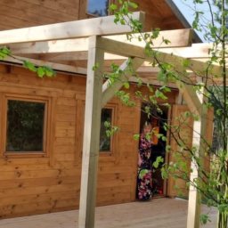 WoodBuild - Drewniany taras z niezadaszoną konstrukcją pergoli w trakcie budowy, drewniany domek w tle, kobieta w kwiecistej sukience w drzwiach domku.