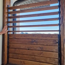 WoodBuild - Drewniana konstrukcja z regulowanymi lamelami i pełnym panelem na dole, widziana z bliska, z dłonią dotykającą ramy, z widokiem na niebo i dachy budynków w tle.