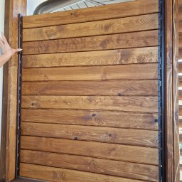WoodBuild - Drewniana żaluzja z poziomych desek w brązowej ramie, zamontowana na zewnątrz budynku, z widoczną ręką kobiety, która ją przesuwa.