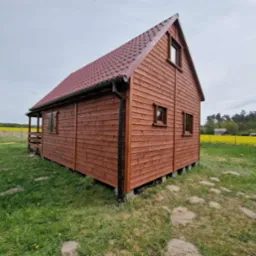 Drewniany domek letniskowy z brązowym dachem i widoczną konstrukcją nośną, otoczony zielenią i polem rzepaku w tle, prezentowany w słoneczny dzień.