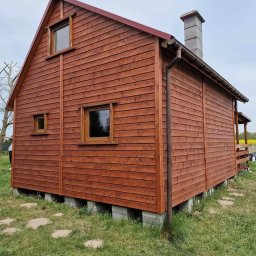 WoodBuild - Nowy, brązowy dom z drewnianą elewacją, widoczny z boku, z betonowym kominem i małymi oknami. Dom stoi na betonowych bloczkach, otoczony trawą i polami.