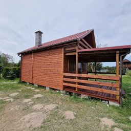 WoodBuild - Drewniany domek z zadaszonym tarasem, wykończony w ciepłym, brązowym odcieniu. Widoczny komin i starannie wykonana konstrukcja. Tło stanowi zieleń trawy i inne zabudowania.