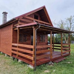 WoodBuild - Czerwony drewniany domek z zadaszonym tarasem i okiennicami, otoczony zieloną trawą. Widoczny komin i elementy konstrukcyjne.