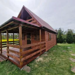 Drewniana altana z zadaszonym tarasem, brązowe deski, czerwony dach, tło: pole kwitnących na żółto kwiatów, widok z boku na tle zielonego trawnika.