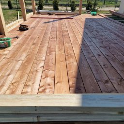 WoodBuild - Montaż desek na tarasie. Widoczny fragment konstrukcji i narzędzia. Część desek ułożona, część jeszcze nie. W tle zieleń i ogrodzenie.