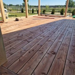 WoodBuild - Drewniany taras w trakcie budowy z widoczną konstrukcją szkieletową i deskami podłogowymi w odcieniach brązu, na tle zieleni i błękitnego nieba.