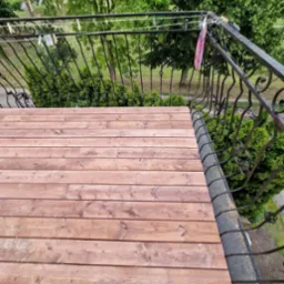 Wykończony taras z desek w odcieniach brązu, z ozdobną, kutą balustradą. Widok z góry na drewnianą podłogę i zieleń w tle. Na balustradzie wiszą pranie i klamerki.