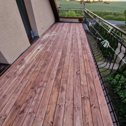 WoodBuild - Balkon z drewnianą podłogą w odcieniach brązu, z metalową balustradą. W tle widok na pole i turbiny wiatrowe. Na balustradzie wiszą klamerki.