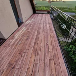 Balkon z drewnianą podłogą w odcieniach brązu, z metalową balustradą. W tle widok na pole i turbiny wiatrowe. Na balustradzie wiszą klamerki.