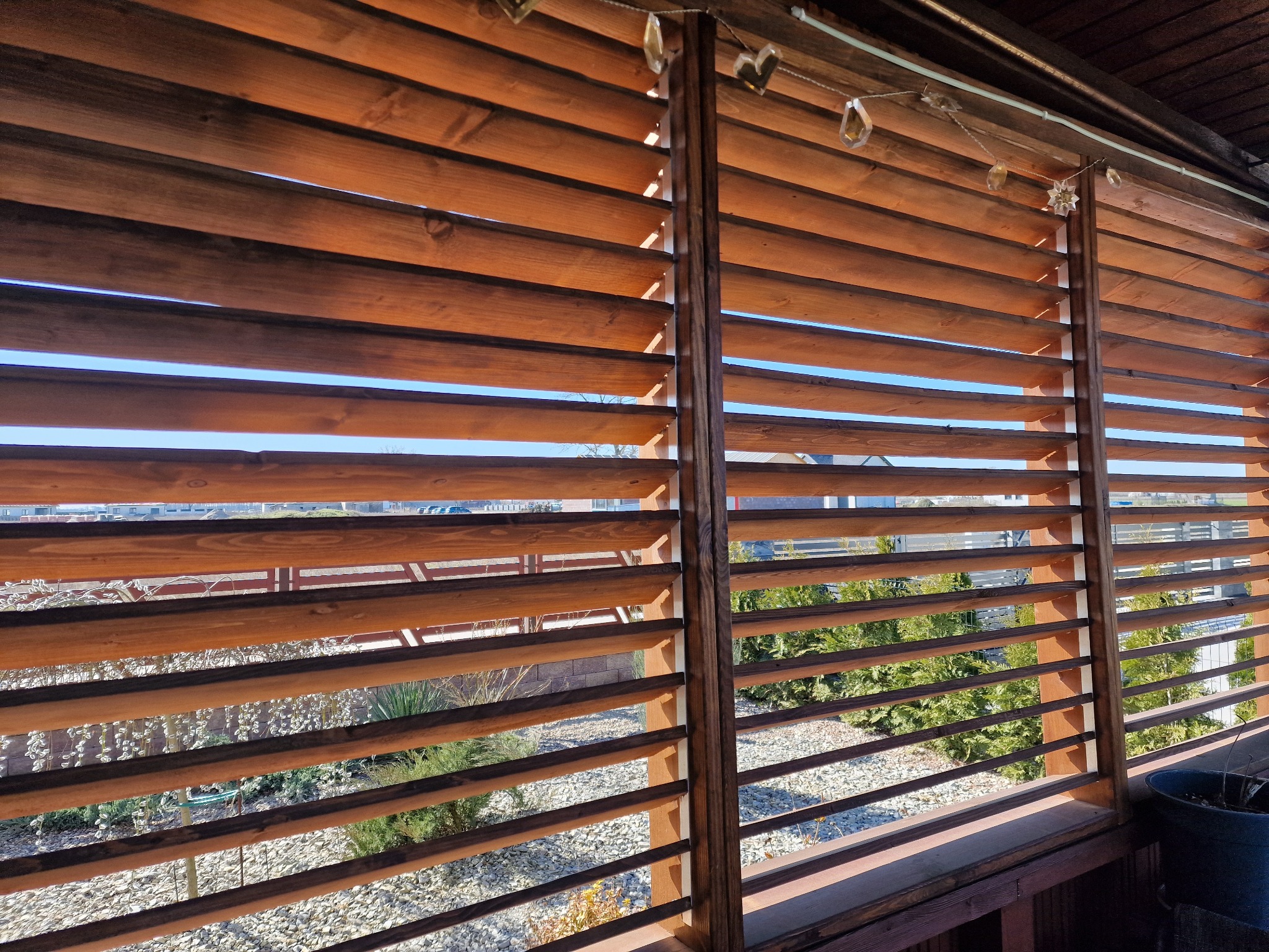 Drewniane żaluzje w brązowym odcieniu, zamontowane w konstrukcji z drewna, przez które widać ogród z kamieniami i roślinami oraz błękitne niebo w oddali. Dekoracyjne lampki wiszą u góry.