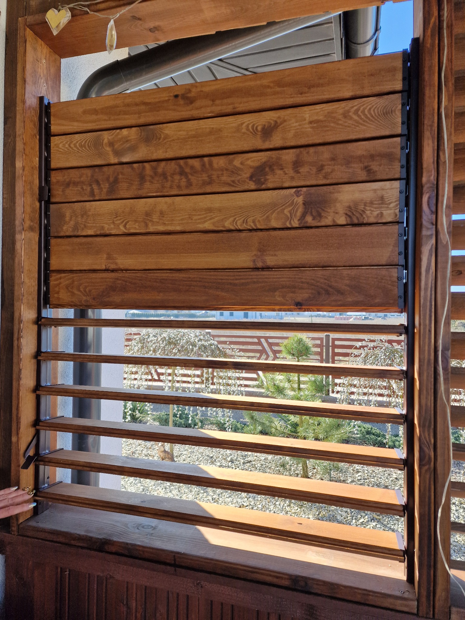 Drewniane żaluzje zewnętrzne z regulowanymi lamelami, widok na ogród z krzewami i drzewami, brązowe drewno i czarne metalowe okucia.