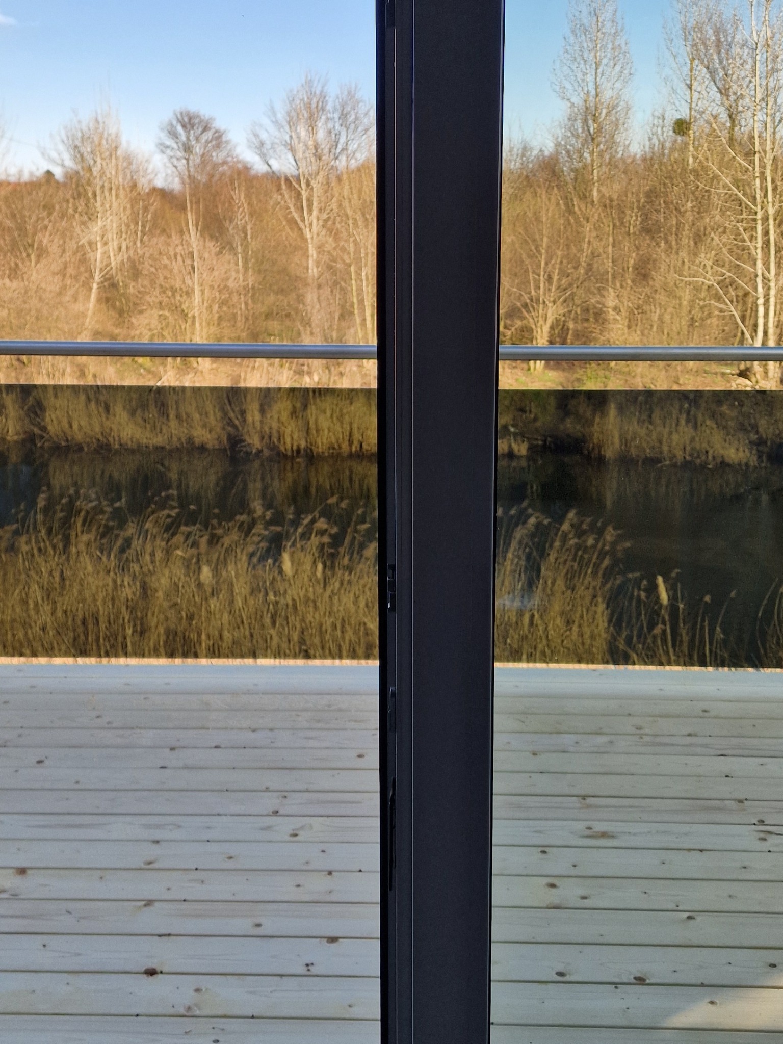 Nowoczesny taras z drewnianą podłogą i szklaną balustradą, widok na jezioro z trzcinami i drzewami w tle, przez przesuwne drzwi.