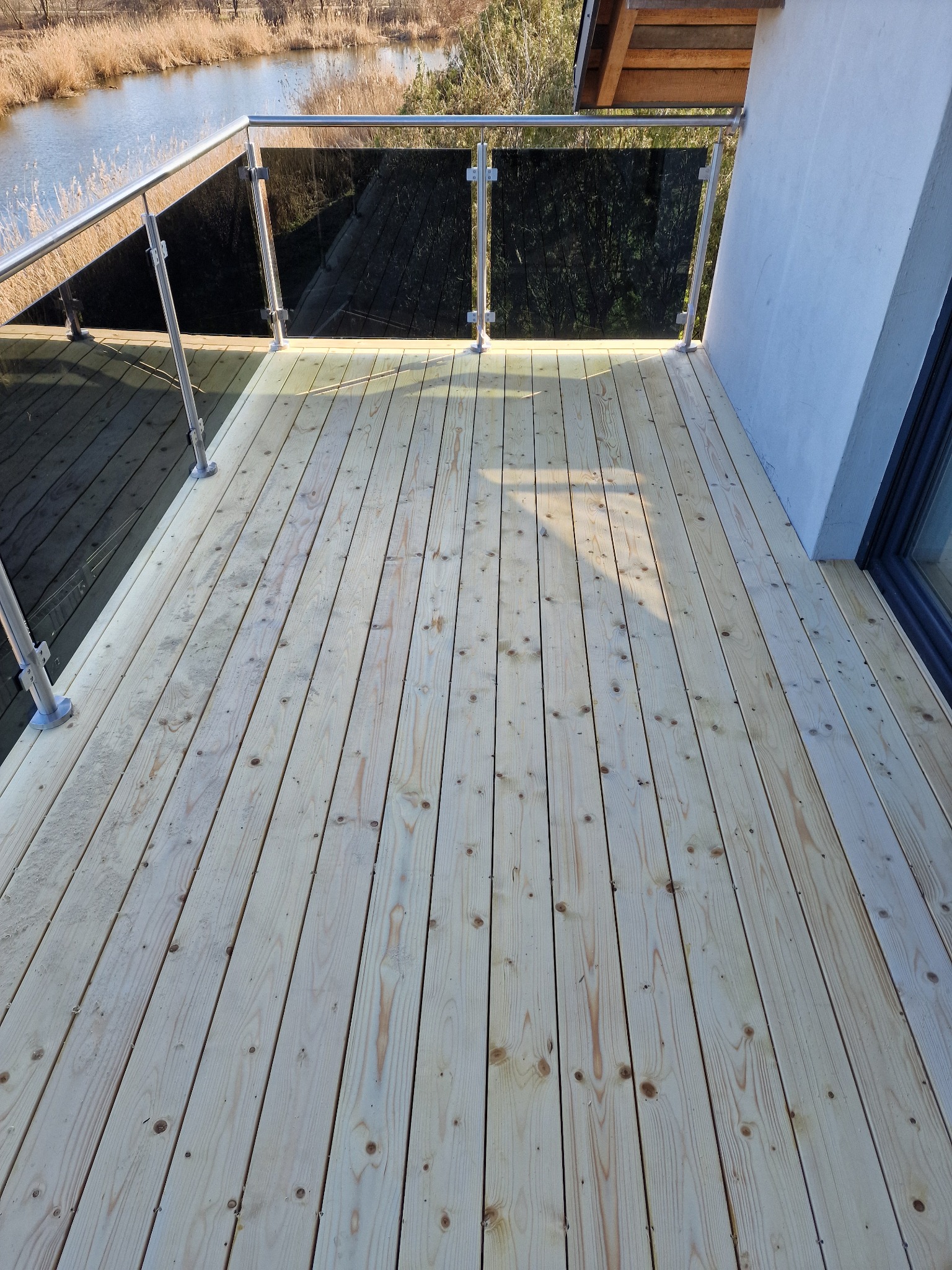 Nowoczesny taras z jasnego drewna z balustradą ze szkła i stali nierdzewnej, widok na jezioro z trzcinami w tle.