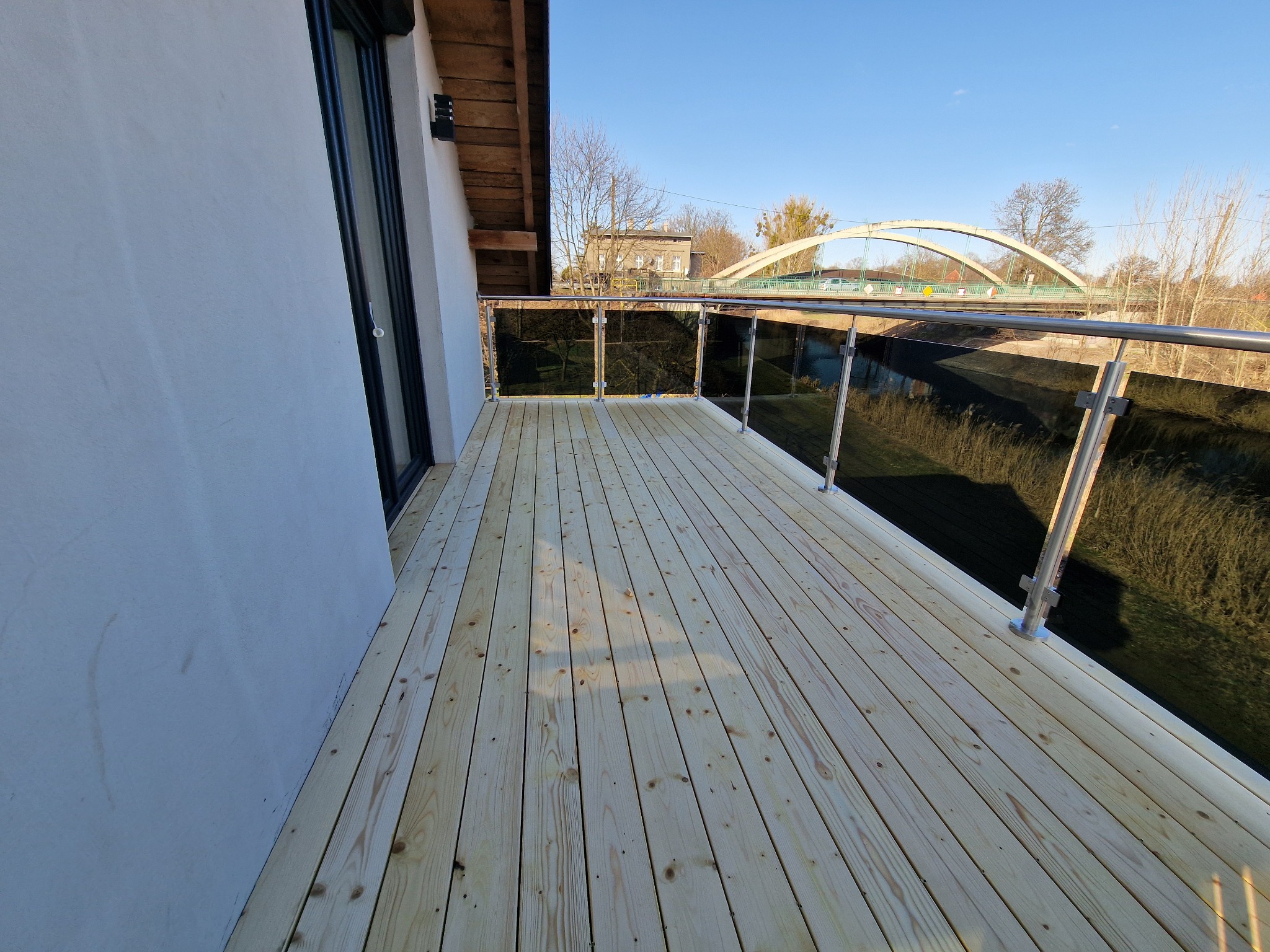 Nowoczesny taras z jasnego drewna z szklaną balustradą, widok na rzekę i most w tle, słoneczny dzień.