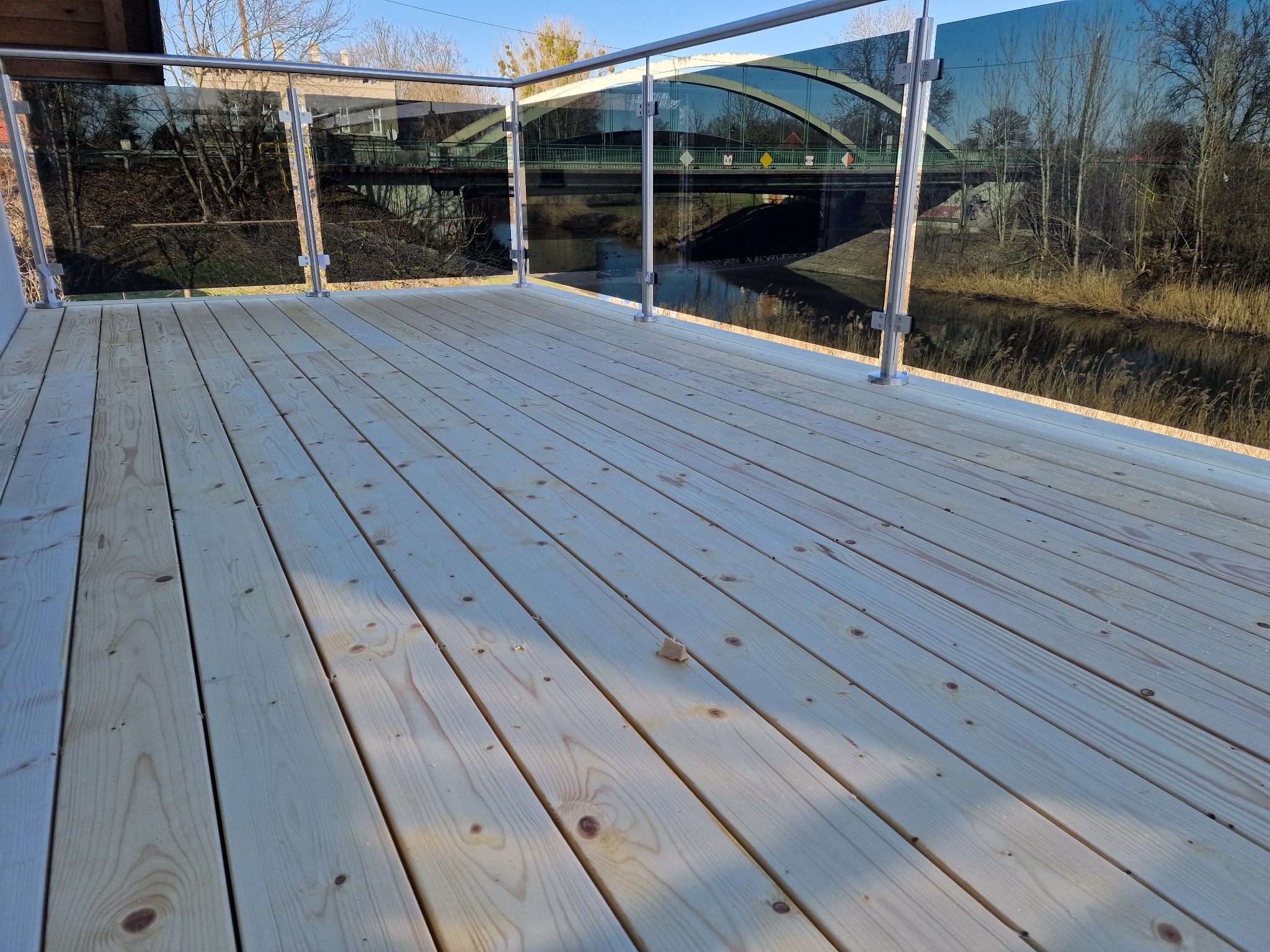 Nowoczesny taras drewniany z balustradą szklaną w Inowrocławiu, z widokiem na rzekę i most w tle.