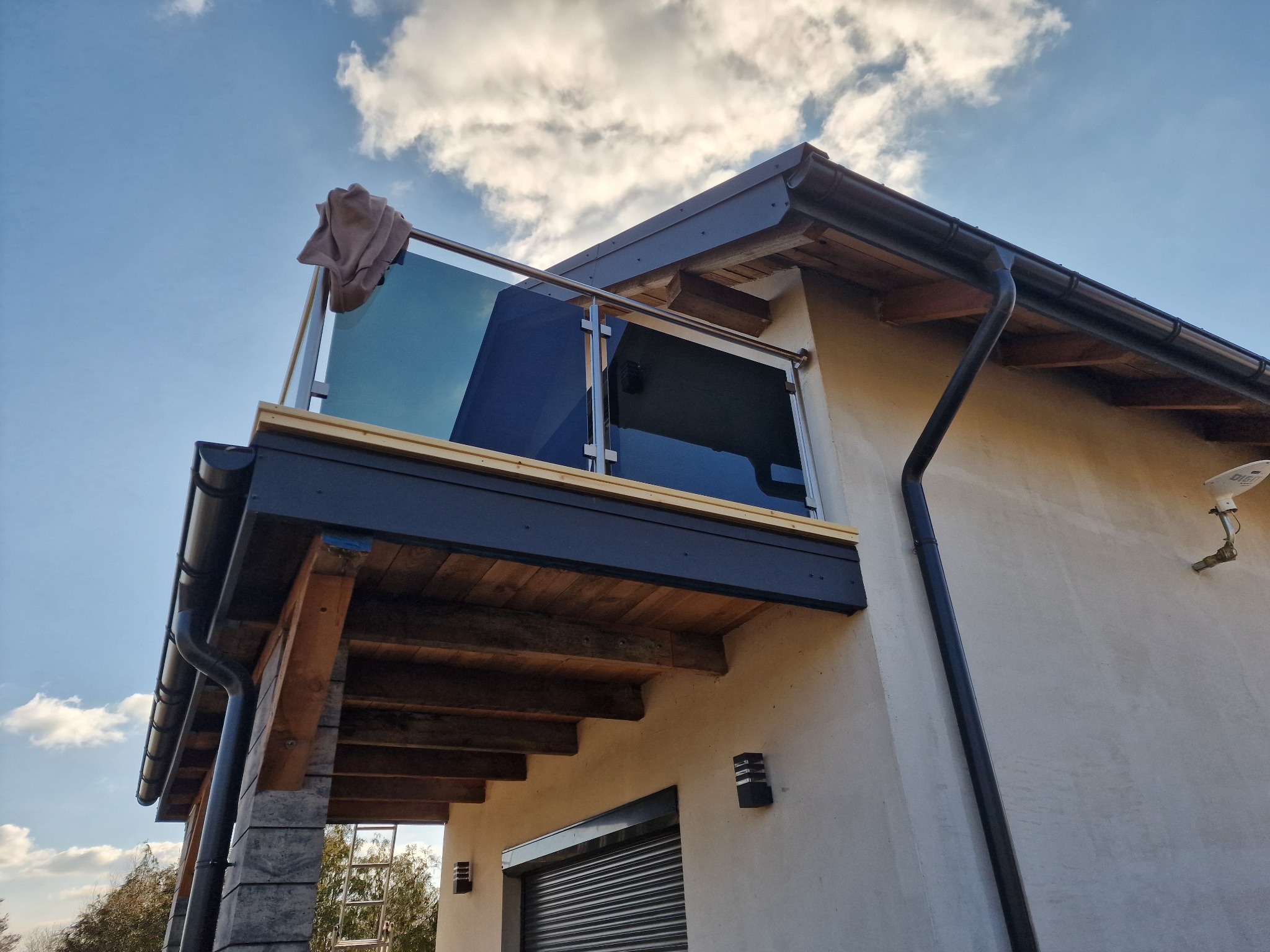 Zewnętrzny widok balkonu z szklaną balustradą i metalową ramą, widoczny fragment elewacji budynku, rynna i fragment nieba z chmurami.