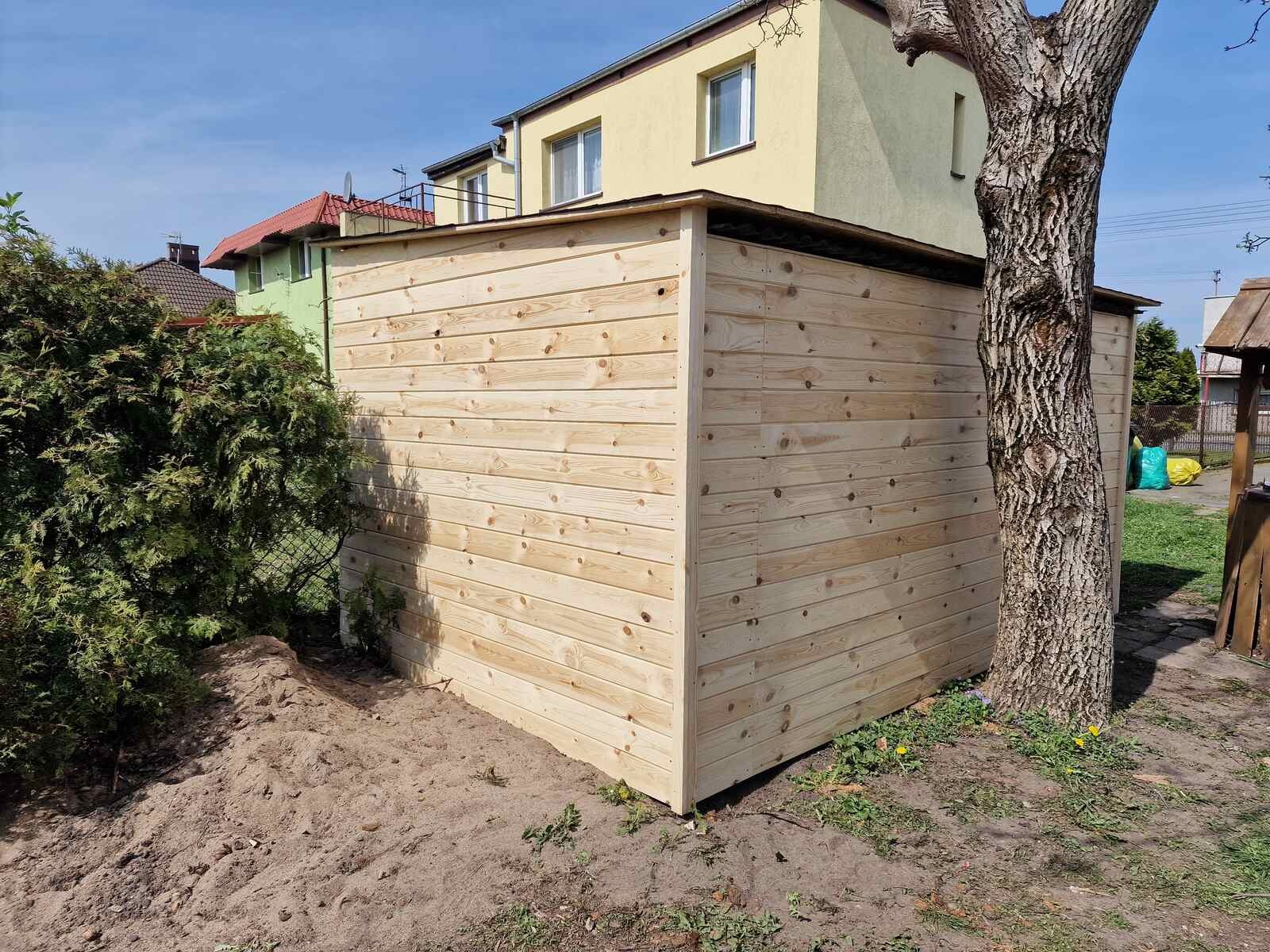 Nowa, surowa, drewniana szopa ogrodowa z desek na tle domu i drzewa, widoczna z boku. Jasne drewno, świeżo zbudowana konstrukcja.