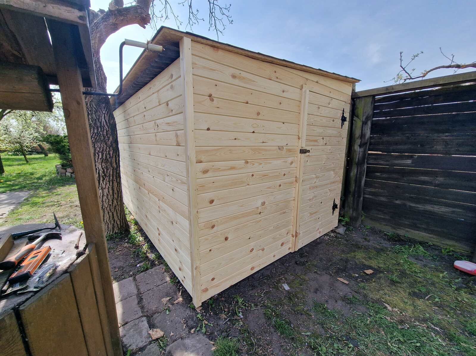 Świeżo postawiona, drewniana, jasna szopa ogrodowa z drzwiami, z czarnymi zawiasami, stojąca na trawie, przy drzewie i płocie. Widoczne narzędzia na stole obok.