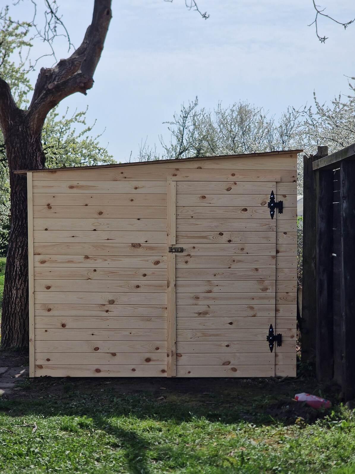 Drewniana szopa narzędziowa z skośnym dachem, umieszczona obok drzewa i ogrodzenia, na tle zielonej trawy i kwitnących drzew. Widoczne zawiasy i zamek.