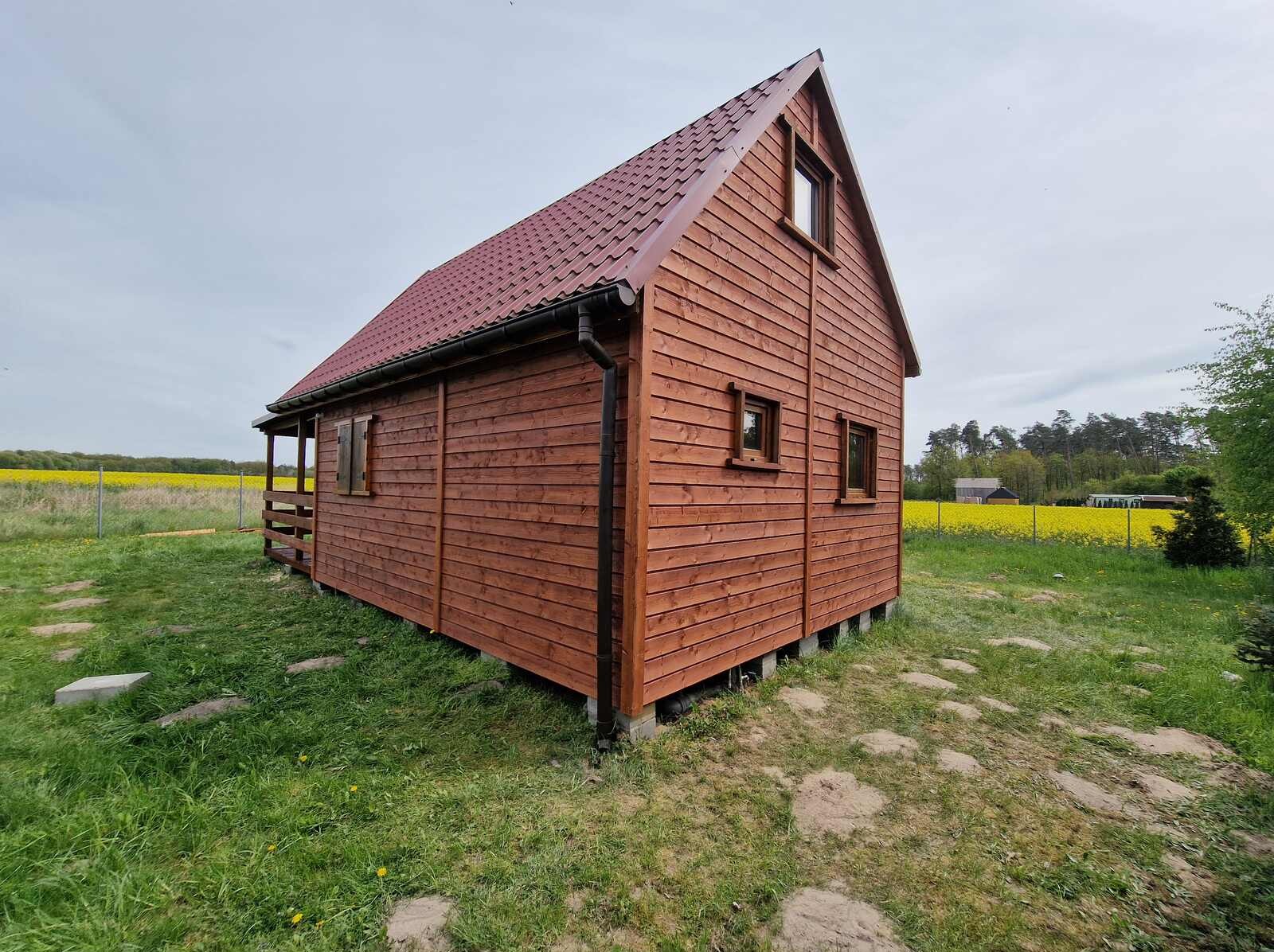 Drewniany domek letniskowy z brązowym dachem i widoczną konstrukcją nośną, otoczony zielenią i polem rzepaku w tle, prezentowany w słoneczny dzień.