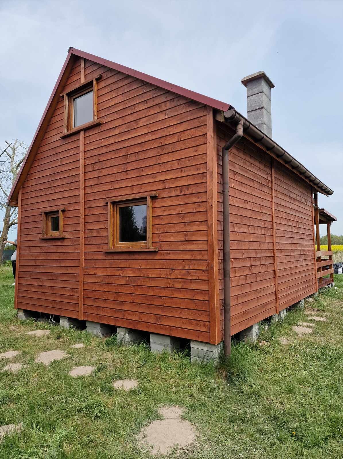 Nowy, brązowy dom z drewnianą elewacją, widoczny z boku, z betonowym kominem i małymi oknami. Dom stoi na betonowych bloczkach, otoczony trawą i polami.