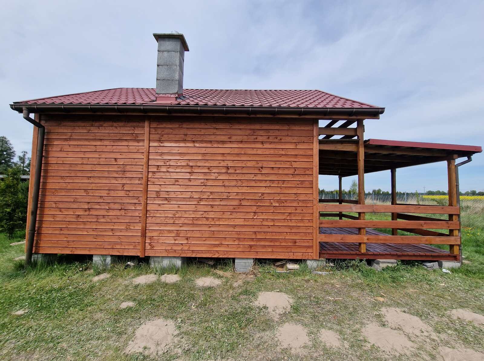 Zewnętrzny widok drewnianej altany z kominem i zadaszonym tarasem, wykończonej czerwoną dachówką, na tle łąki i błękitnego nieba.