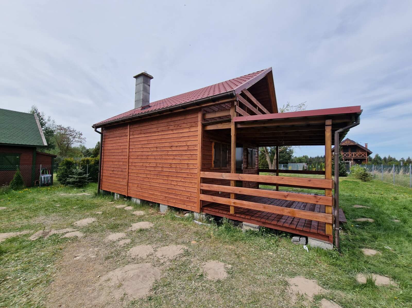 Drewniany domek z zadaszonym tarasem, wykończony w ciepłym, brązowym odcieniu. Widoczny komin i starannie wykonana konstrukcja. Tło stanowi zieleń trawy i inne zabudowania.