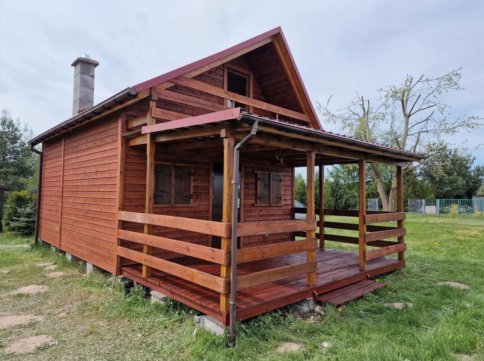 Czerwony drewniany domek z zadaszonym tarasem i okiennicami, otoczony zieloną trawą. Widoczny komin i elementy konstrukcyjne.