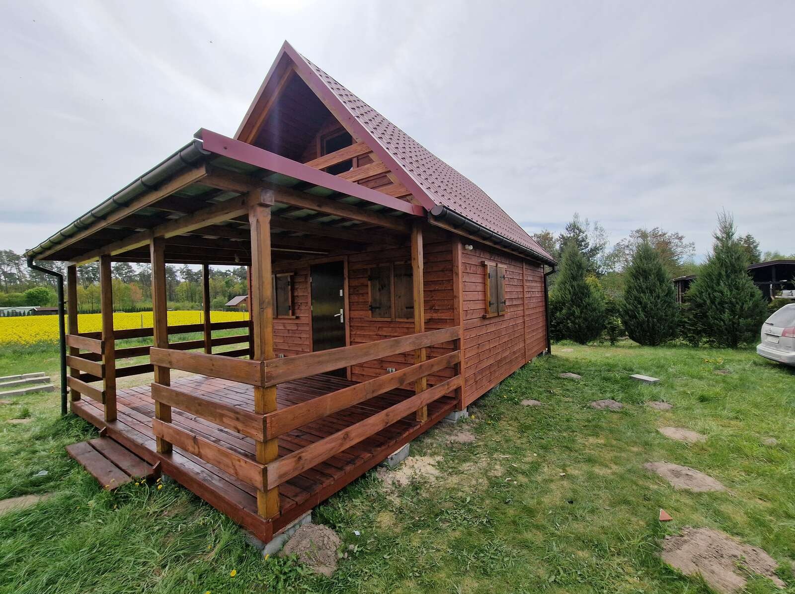 Drewniana altana z zadaszonym tarasem, brązowe deski, czerwony dach, tło: pole kwitnących na żółto kwiatów, widok z boku na tle zielonego trawnika.