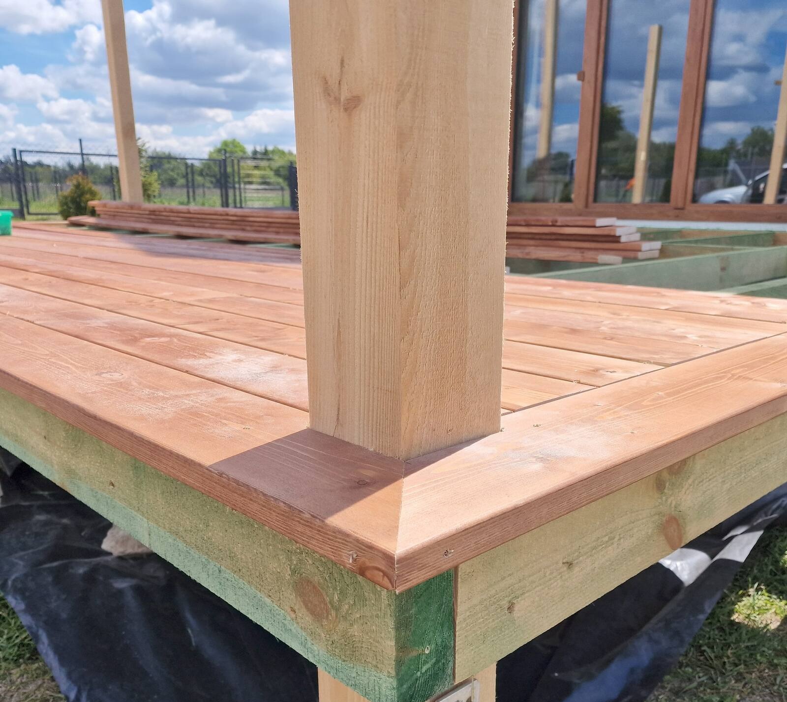 Fragment konstrukcji tarasu drewnianego w trakcie budowy, widoczne deski podłogowe, słup podporowy i elementy ramy, tło z zielenią i niebem.