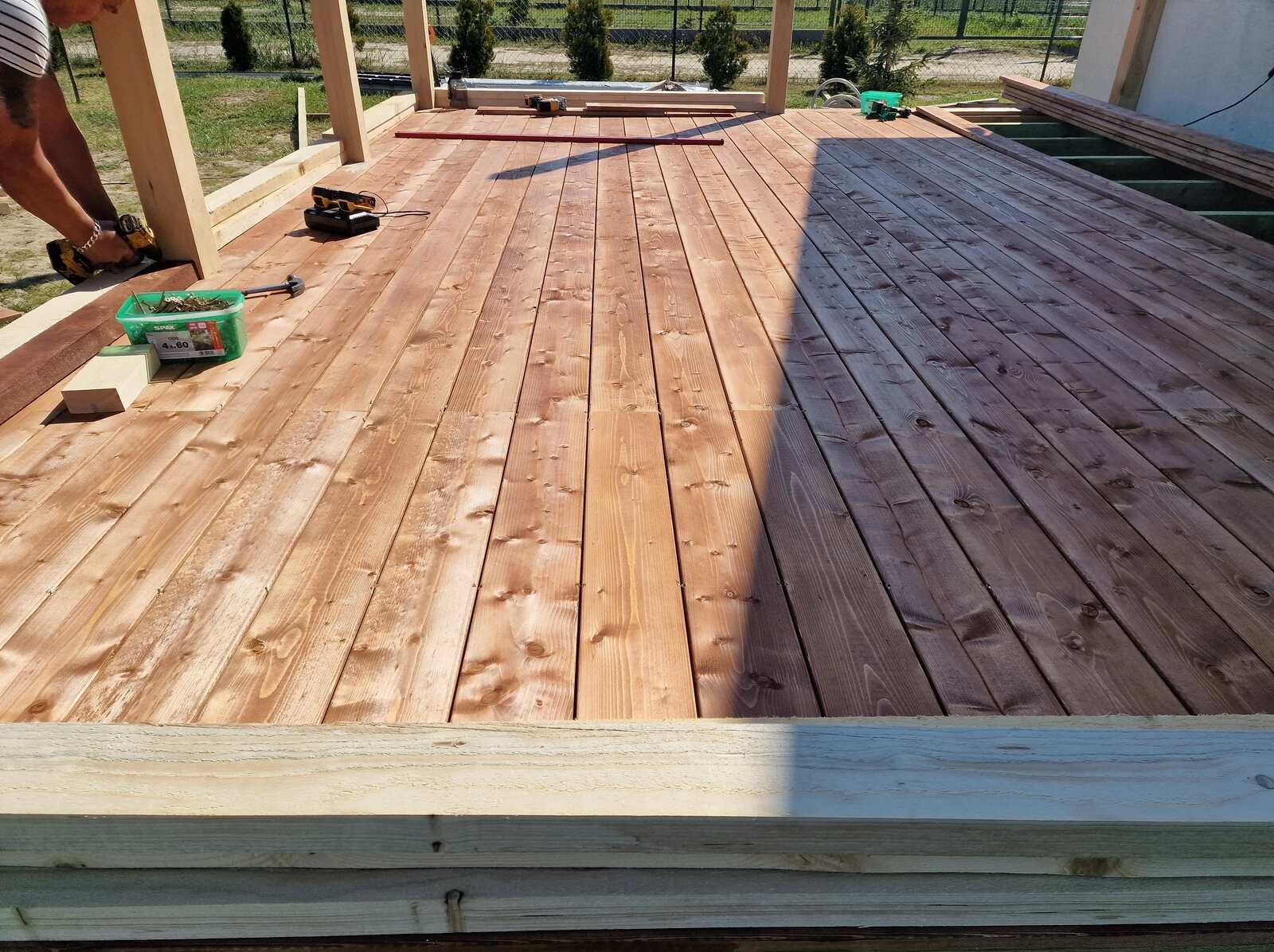 Montaż desek na tarasie. Widoczny fragment konstrukcji i narzędzia. Część desek ułożona, część jeszcze nie. W tle zieleń i ogrodzenie.