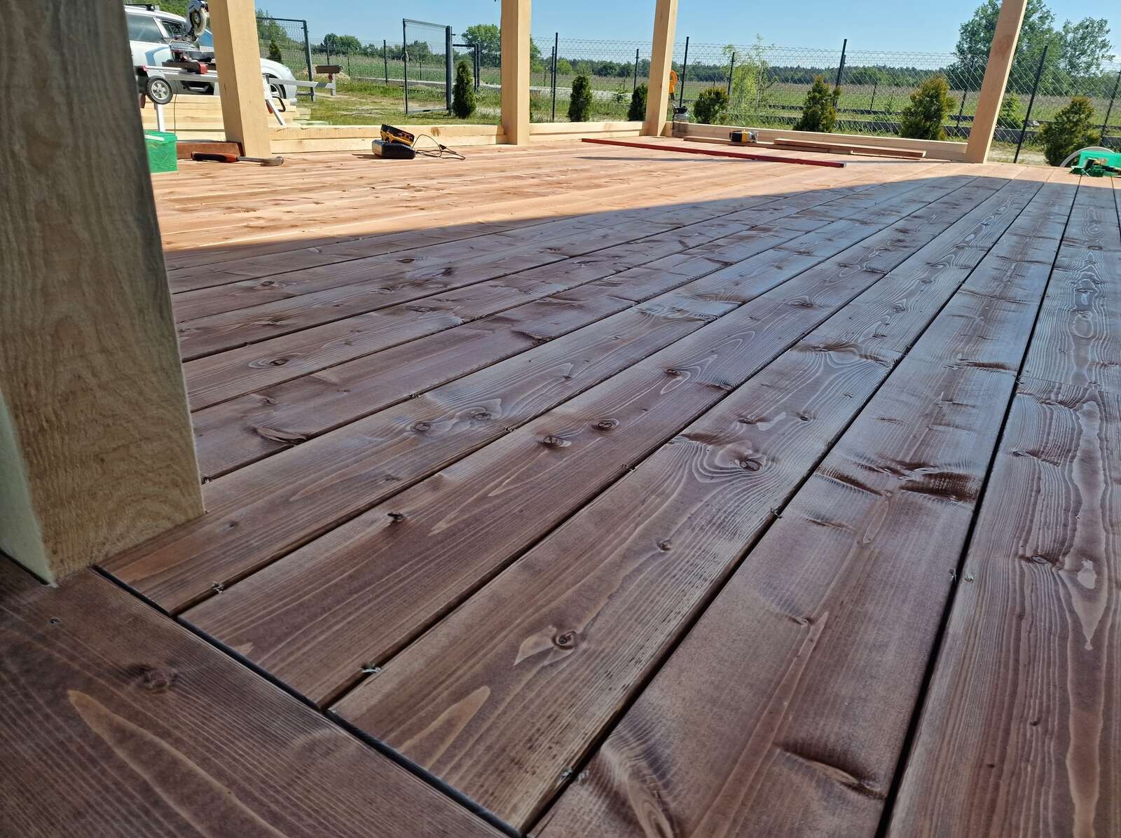 Brązowe deski tarasowe ułożone pod lekkim kątem, z widocznymi słupami konstrukcji i narzędziami w tle, sugerujące świeżo zakończone prace.