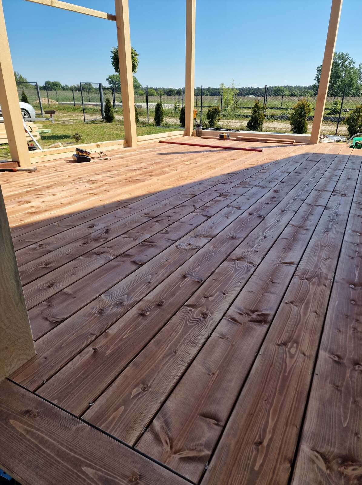 Drewniany taras w trakcie budowy z widoczną konstrukcją szkieletową i deskami podłogowymi w odcieniach brązu, na tle zieleni i błękitnego nieba.