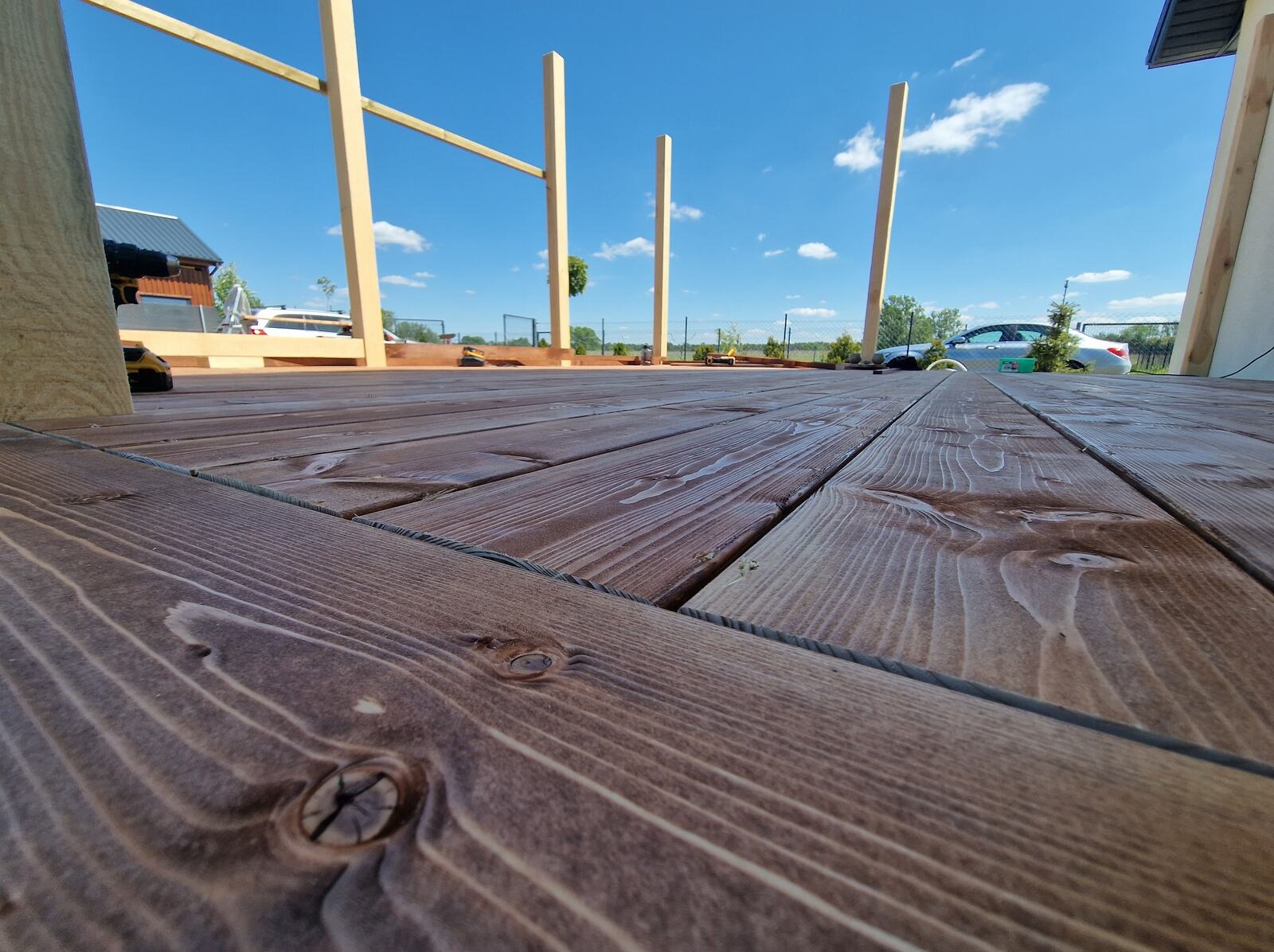 Montaż brązowego tarasu z desek z widokiem na niebo i elementy konstrukcyjne pergoli w tle. Ujęcie z dołu podkreśla strukturę drewna i głębię przestrzeni.
