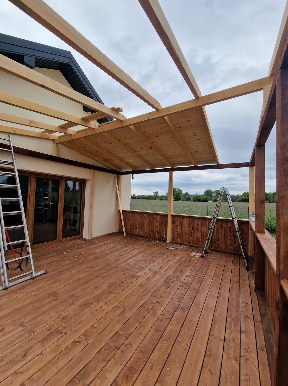 Drewniany taras z pergolą w trakcie budowy. Widoczne deski tarasowe, konstrukcja dachu i balustrady. Dwie drabiny opierają się o konstrukcję. Tło stanowi zielone pole.