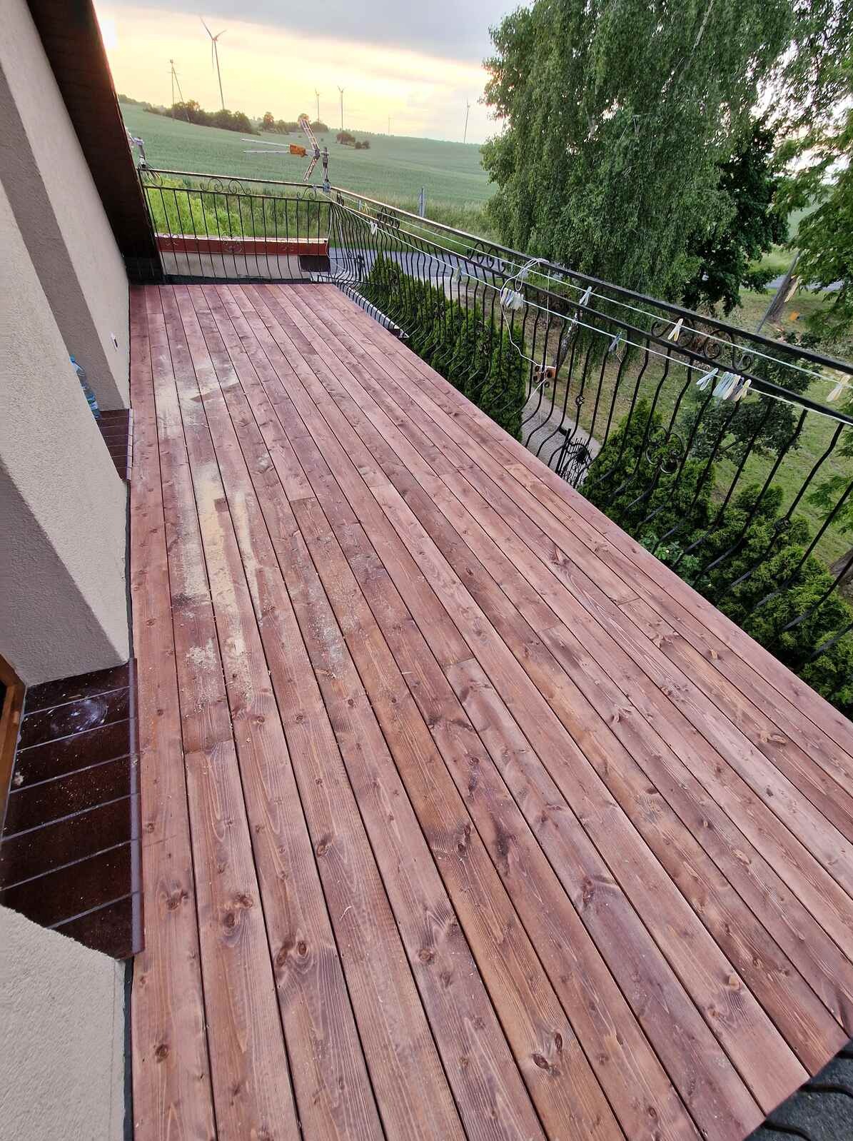 Wykończony taras z desek z metalową balustradą. Widok na zielone pola i turbiny wiatrowe w oddali. Częściowo widoczne pranie na balustradzie.