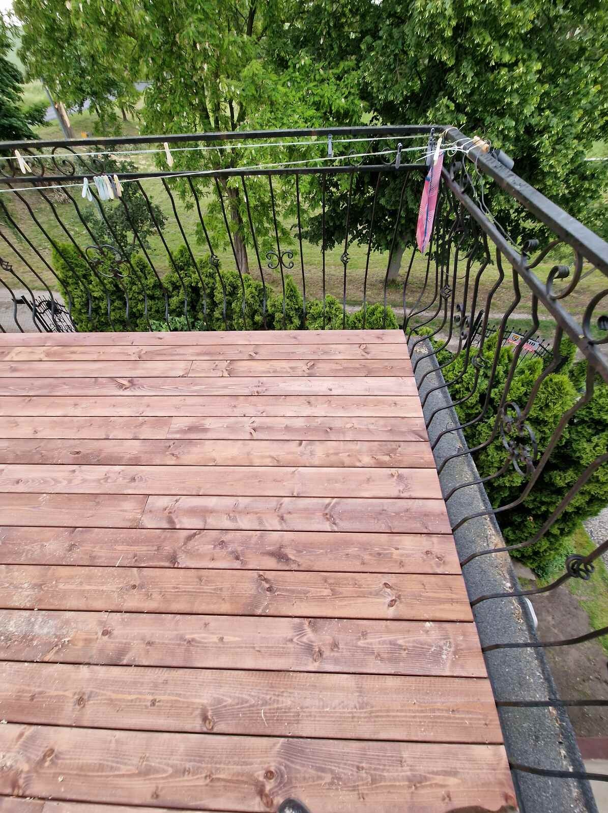 Wykończony taras z desek w odcieniach brązu, z ozdobną, kutą balustradą. Widok z góry na drewnianą podłogę i zieleń w tle. Na balustradzie wiszą pranie i klamerki.