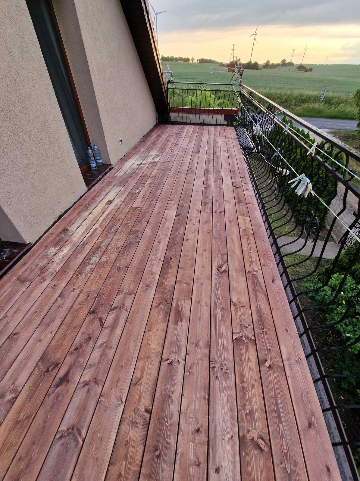 Balkon z drewnianą podłogą w odcieniach brązu, z metalową balustradą. W tle widok na pole i turbiny wiatrowe. Na balustradzie wiszą klamerki.