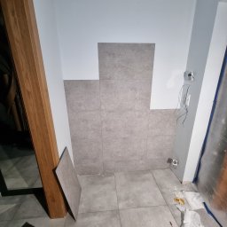 MAŁY - Fragment ściany łazienki w trakcie układania szarych, kwadratowych płytek ceramicznych, widoczne wystające przewody elektryczne i narzędzia na podłodze.