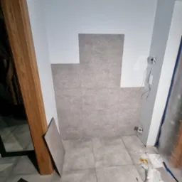 Fragment ściany łazienki w trakcie układania szarych, kwadratowych płytek ceramicznych, widoczne wystające przewody elektryczne i narzędzia na podłodze.