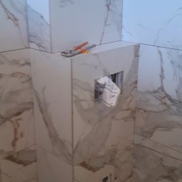 MAŁY - Łazienka w trakcie wykańczania: obudowa stelaża WC z płytkami imitującymi marmur, widoczne narzędzia, rura kanalizacyjna i otwór na przycisk spłukujący.