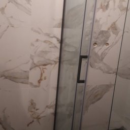 MAŁY - Szklana kabina prysznicowa z czarnym uchwytem na tle marmurowych płytek ściennych i podłogi z czarnego kamienia z złotymi żyłami.